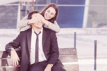 Closeup portrait of joyful middle-aged business woman covering eyes of her male colleague outdoors. Man is sitting on bench. Woman is standing behind.の写真素材