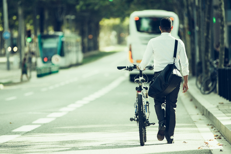 Back view of businessman walking with shoulder bag and bike in street. Young male office employee going after work. Lifestyle conceptの写真素材