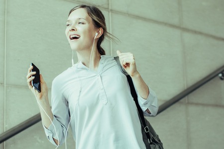 Excited carefree female intern listening to music and dancing outdoors. Cheerful student girl full...
