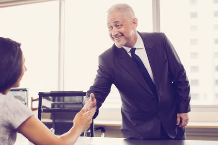 Closeup portrait of smiling senior business man standing and extending arm to blurred business woman for handshake with office window in background. Woman is sitting back to camera.の写真素材