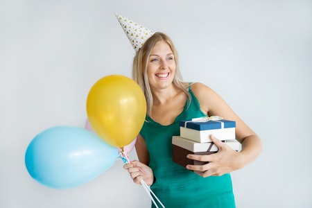 Overjoyed beautiful girl enjoying birthday party. Happy blonde young Caucasian woman in festive hat holding air balloons and embracing gifts. Festive party conceptの写真素材