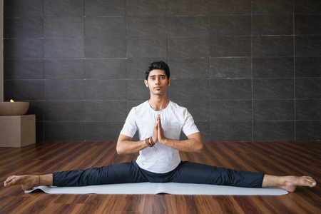 Experienced Indian yogi doing splits in gym. Man looking at camera, holding hands together and practicing yoga. Yogi concept. Front view.の写真素材