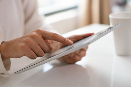Cropped view of freelancer working with touchpad at home. Woman using tablet at white desk with cup of tea. Mobile computer conceptの写真素材
