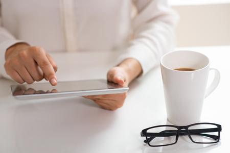 Professional still working during coffee break. Closeup of female employee using tablet at white desk with cup of coffee and glasses. Coffee break conceptの写真素材