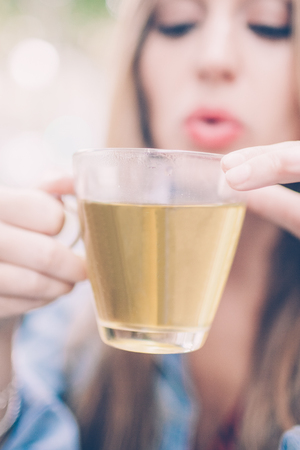 Closeup portrait of serious young beautiful fair-haired woman holding cup of tea and blowing on it outdoors in summer with focus on cupの写真素材
