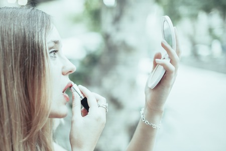 Closeup portrait of young beautiful fair-haired woman holding mirror and rouging lips outdoors in summer. Side view.の写真素材