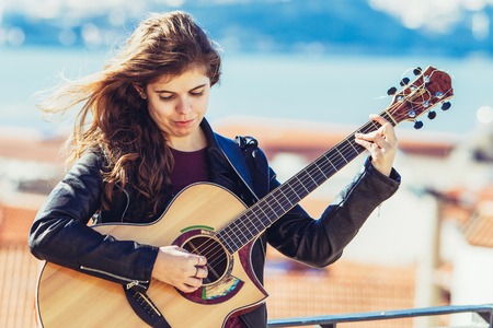 Closeup portrait of serious young beautiful woman standing on rooftop in city and playing guitar. Front view.の写真素材
