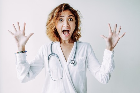 Portrait of scared or excited young Asian female doctor wearing uniform and stethoscope looking at camera and screaming. Excitement or fear conceptの写真素材
