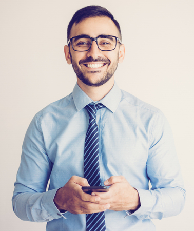 Closeup portrait of smiling young handsome business man looking at camera, holding smartphone and texting on it. Technology concept. Isolated front view on grey background.の写真素材