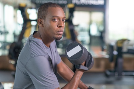 Black man training with dumbbell and looking at camera. Young guy wearing t-shirt and sitting in gym...
