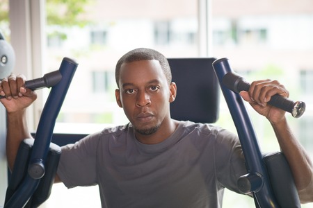 Black man training on pec deck fly machine and looking at camera. Young guy wearing t-shirt with...