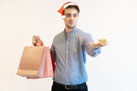Content Christmas shopper giving little gift. Handsome young man in Santa cap holding shopping bags and showing present box on palm. Present buying conceptの写真素材
