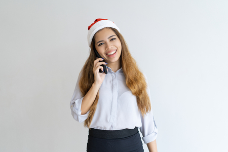 Smiling woman talking on smartphone and wearing Santa Claus hat. Pretty lady looking at camera. Christmas and communication concept. Isolated front view on white background.の写真素材