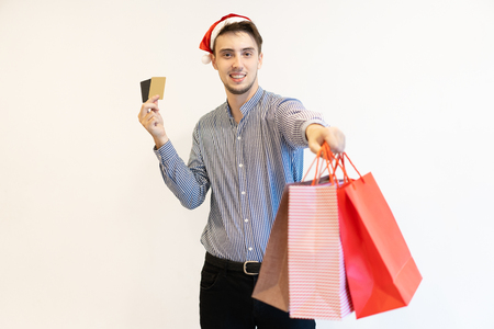 Happy cardholder recommending Christmas shopping. Handsome young man in Santa hat holding credit cards and giving shopping bags at camera. Christmas sale conceptの写真素材