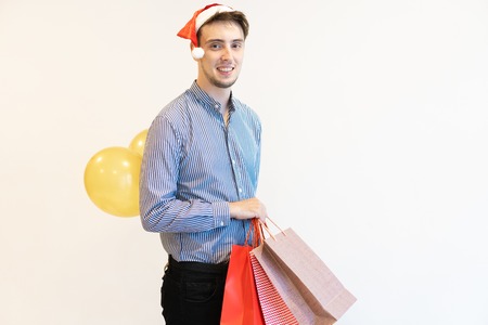 Smiling guy planning Christmas surprise. Handsome young man in Santa cap carrying shopping bags and...