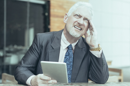 Closeup portrait of pensive senior handsome man looking upwards, using tablet computer and sitting at table in outdoor cafe. Front view.の写真素材