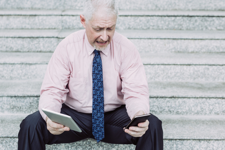 Closeup portrait of worried senior handsome man using tablet computer and smartphone while sitting...