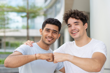 Portrait of happy multiethnic male friends making fist bump. Young Indian and Hispanic male students embracing, looking at camera and smiling outdoors. Male friendship conceptの写真素材