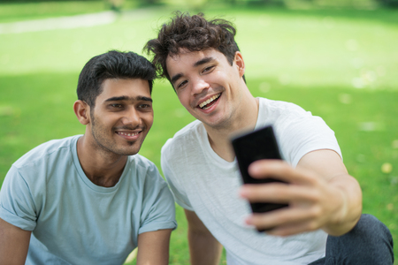 Cheerful carefree young men taking selfie on phone while enjoying free time in park. Excited friends in casual clothing posing for photo on smartphone. Friendship or Selfie conceptの写真素材