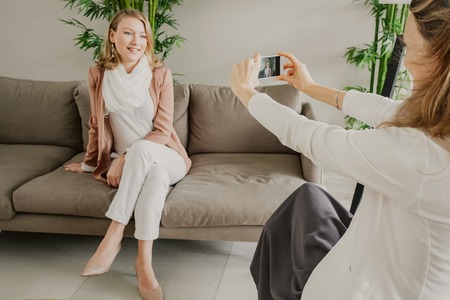 Woman taking photo of her beautiful female friend who is sitting on couch in lounge with wall and bamboo in background. Women friendship concept.の写真素材