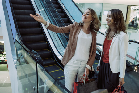 Closeup portrait of two smiling beautiful women holding paper-bags in shopping mall with escalator in background. One woman is pointing at something. Shopping concept.の写真素材