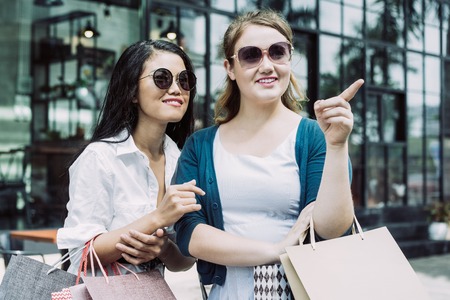 Closeup portrait of two smiling young beautiful multiethnic women shopping and holding paper-bags in city with building in background. One woman is pointing at something. Shopping concept. Front view.の写真素材