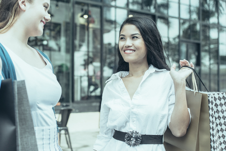 Closeup portrait of two smiling young beautiful multiethnic women chatting and holding paper-bags in city with building in background. Shopping concept.の写真素材