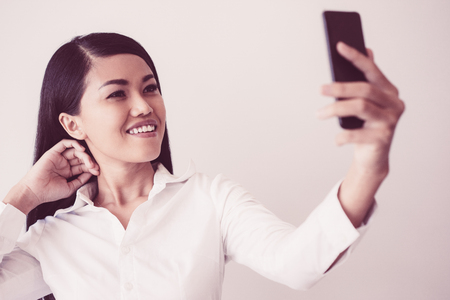 Closeup portrait of smiling young pretty Asian business woman posing and taking selfie photo on smartphone. Selfie concept. Isolated view on grey background.の写真素材