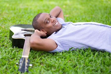 Happy black man lying on grass and guitar in park. Handsome young man looking at camera. Guitarist and rest concept.の写真素材