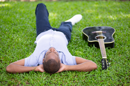 Black man lying upside down on grass and guitar. Handsome young man in park. Guitarist and rest concept.の写真素材