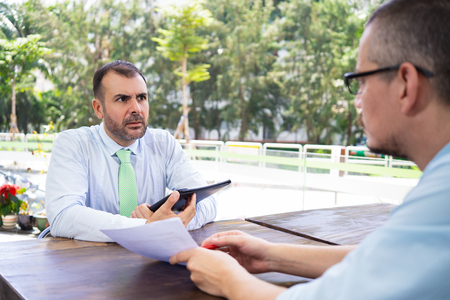 Business consultant explaining papers to Hispanic client. Astonished man with stubble holding folder and attentively listening to advisor at meeting. Examining documents conceptの写真素材