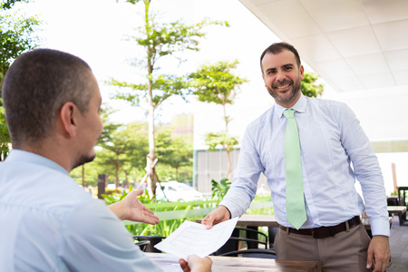 Happy Hispanic manager giving business papers to boss and looking at camera. Cheerful excited man reporting in front of executive in cafe. Business courier conceptの写真素材