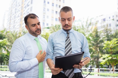 Pensive concentrated colleagues analyzing document while standing in city. Serious busy men working with data together. Examining files conceptの写真素材