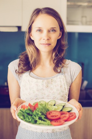 Closeup portrait of content young beautiful woman looking at camera and holding plate with sliced vegetables in kitchen. Healthy cooking concept. Front view.の写真素材