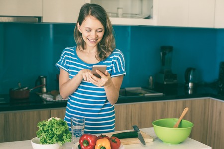 Closeup portrait of smiling young beautiful woman using smartphone, reading recipe and cooking in...
