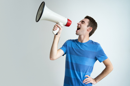 Excited attractive man shouting into megaphone. Young guy keeping hand on hip. Promotion concept. Isolated front view on white background.の写真素材