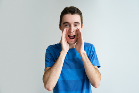 Serious man holding hands near mouth and shouting loud. Young guy looking at camera. Announcement concept. Isolated front view on white background.の写真素材