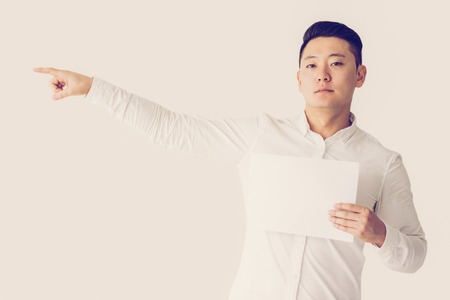 Serious Asian man pointing finger aside and holding empty sheet of paper. Young businessman presenting something on paper. Business and advertising conceptの写真素材