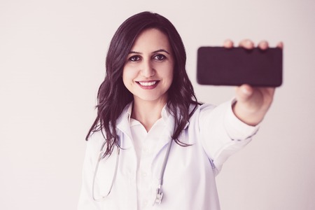 Closeup portrait of smiling beautiful dark-haired female doctor looking at camera and showing smartphone screen.の写真素材