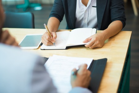 Close-up of woman sitting at table and writing notes in notepad, her male colleague sitting opposite with documents in folder. Education or business conceptの写真素材