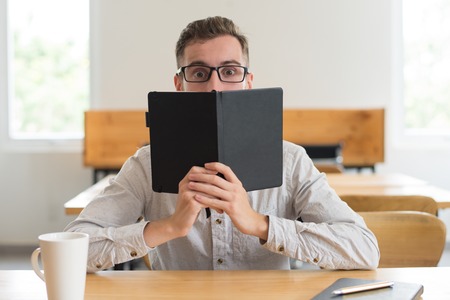 Male student reading book and hiding face behind it at desk in classroom. Playful young man looking...