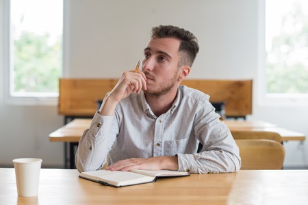 Pensive male student sitting at desk in classroom. Young man reading textbook and thinking. Education and knowledge concept.の写真素材