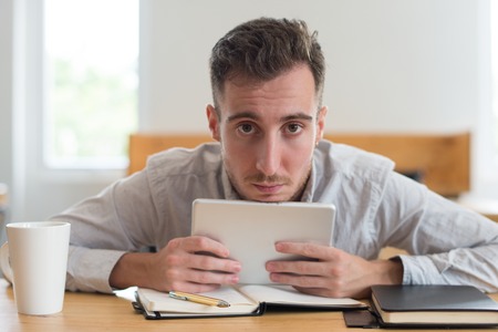 Serious male student holding tablet and looking at camera in classroom. Young man browsing on gadget at desk. Education and technology concept. Front view.の写真素材