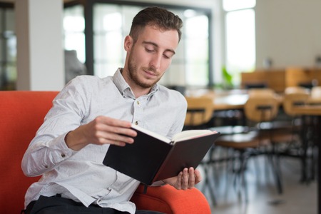 Focused guy reading book on sofa in lobby. Young man studying. Education and literature concept.