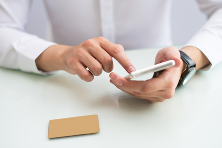 Close-up of unrecognizable entrepreneur checking finances while using online payment app on smartphone. Businessman sitting at table and reading information on phone. Browsing conceptの写真素材