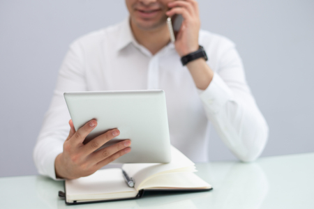Smiling businessman using digital tablet and talking on phone. Young Caucasian businessman sitting at table and networking on touchpad. Technology in business conceptの写真素材