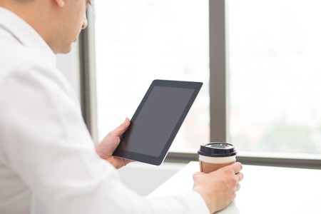 Concentrated businessman reading internet article on tablet and drinking coffee. Serious man sitting at table and working with online data. Connection conceptの写真素材