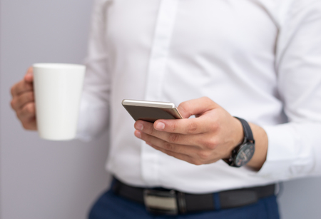 Close-up of young businessman using mobile phone indoors. Caucasian man standing with tea cup and reading or texting message on smartphone. Mobile communication conceptの写真素材