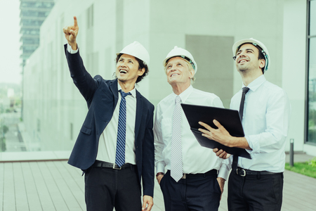 Closeup of three smiling diverse business people wearing helmets, looking at something and standing outdoors with building in background. Engineers concept. Front view.の写真素材