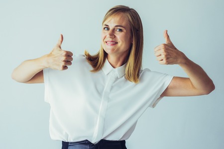Portrait of successful young Caucasian businesswoman wearing white shirt showing thumbs up and...